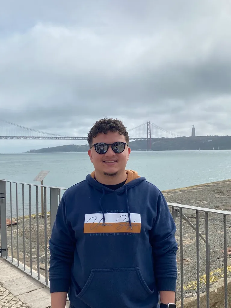 Marcelo Junior, de óculos escuros e moletom azul, com a ponte 25 de Abril e o rio Tejo ao fundo, em Lisboa
